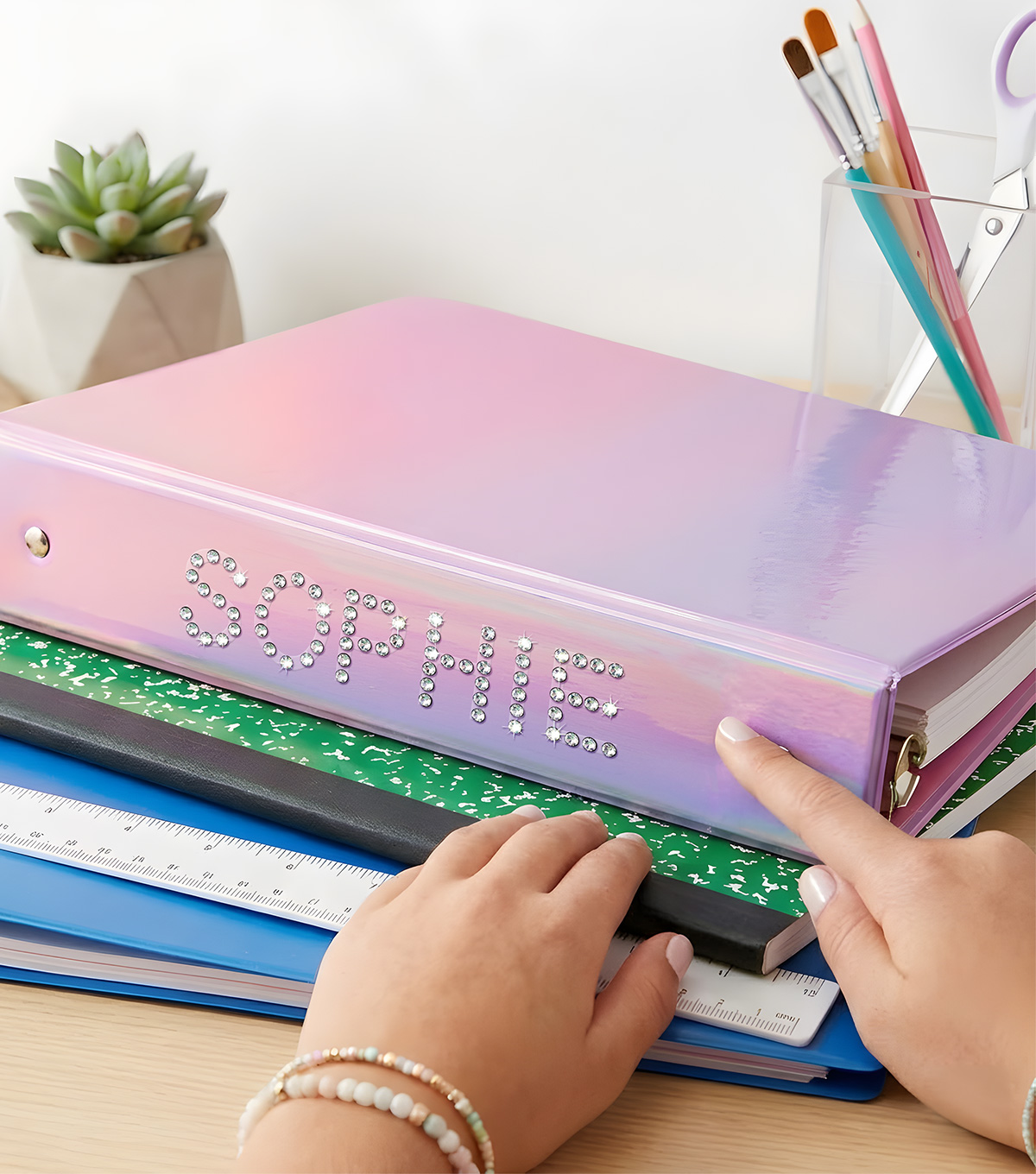 Crystal Stickers rhinestone alphabet letters applied to a school binder to spell the name Sophie, resting on a desk with notebooks and craft supplies.